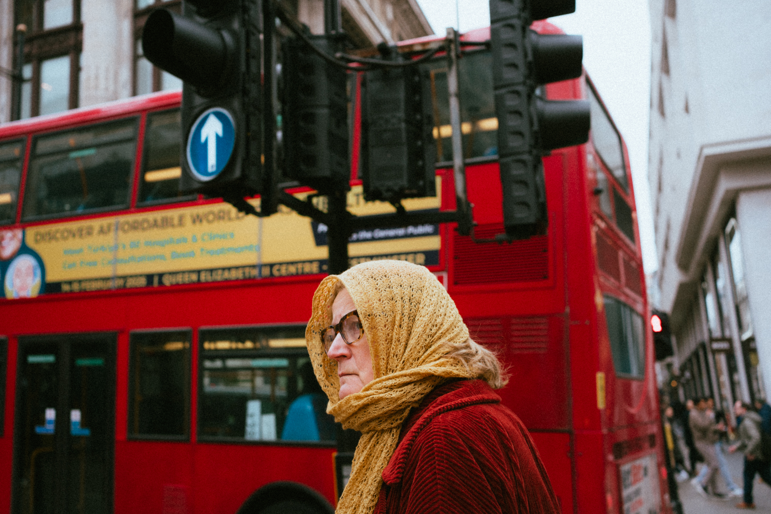 Woman in a red coat and a yellow scarf wrapped around her head passes by a buss with the same two colors. 